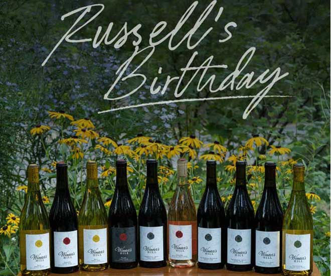 Russell’s birthday sale is May 21st, 2026! - Winter's Hill Estate Row of wine bottles on a wooden table with a handwritten 'Russell's Birthday' sign above, set against yellow flowers and greenery.