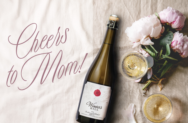 WC-WintersHill-26MothersDay - Winter's Hill Estate Champagne bottle with two glasses and pink flowers on a beige cloth, text reads 'Cheers to Mom!'
